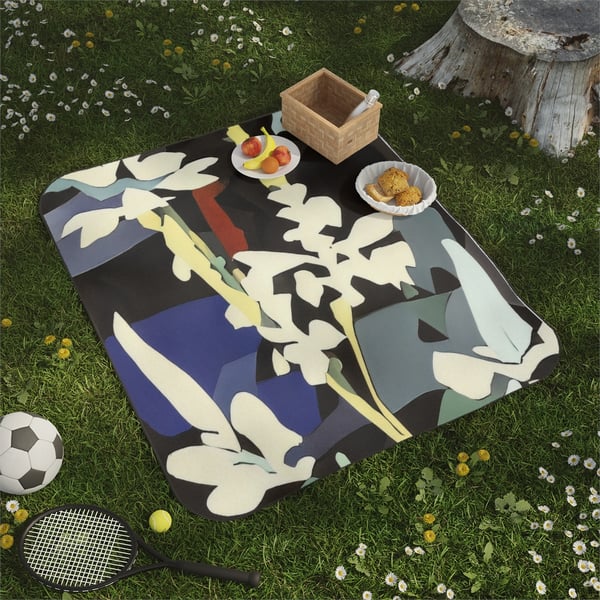 Digital artwork depicting a Montessori-inspired picnic scene with a soft mink polyester picnic blanket featuring a one-sided print of abstract nature shapes, laid out on a grassy field adorned with wildflowers. The blanket's water-resistant oxford cloth backing is not visible, and it is accompanied by a picnic basket, fresh fruit on a plate, a serving of cornbread, a soccer ball, and a tennis racket, emphasizing the product's use for various outdoor activities.