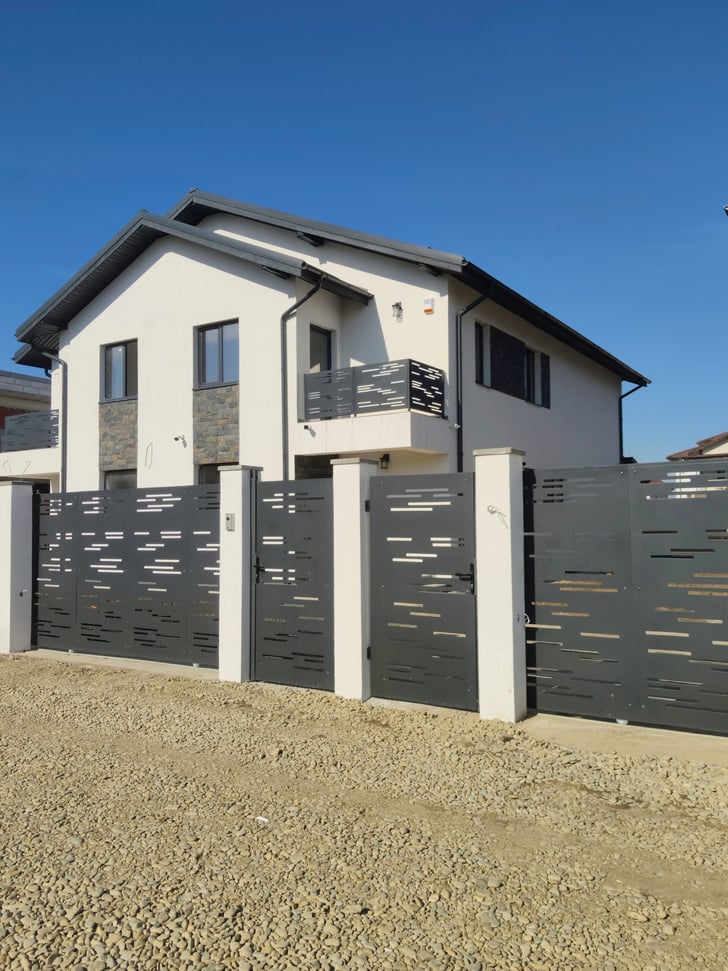 A two-story house with a mix of stone and white stucco exteriors stands under a clear blue sky. The property is surrounded by a modern metal gate with geometric cutouts, and a driveway of small stones leads to the entrance.